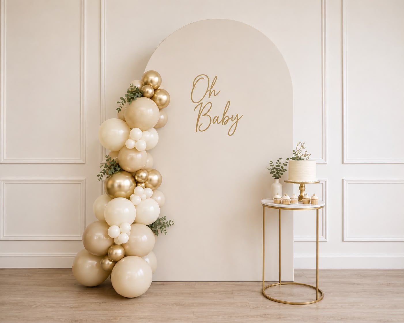 Oh Baby arch backdrop with a nude, cream and gold balloon garland and a small cake table