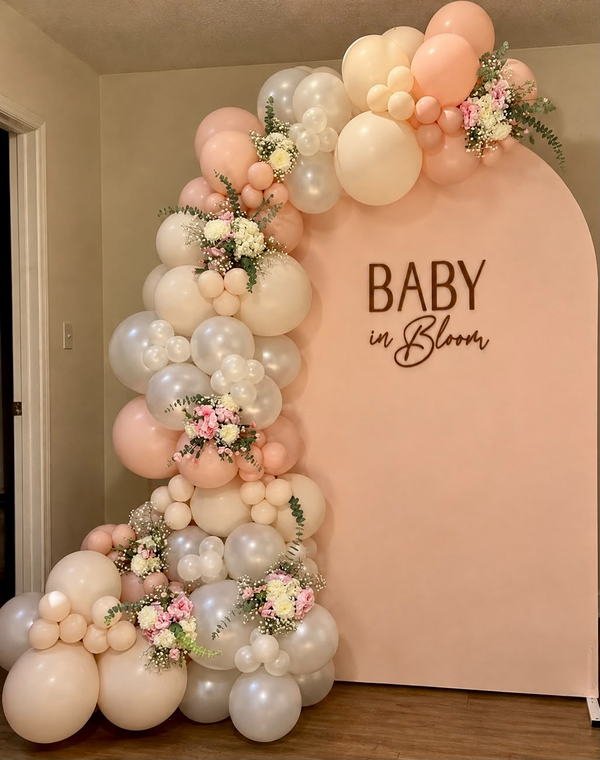 Peach arch backdrop with a Baby in Bloom sign, a peach, cream and white balloon garland and silk florals
