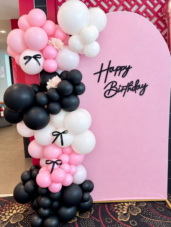 Pink arch backdrop with a Happy Birthday sign and a pink, white and black balloon garland finished with satin bows